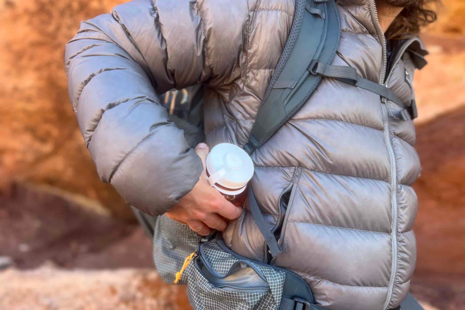 A hiker pulls a water bottle out of a side pocket while wearing a grey backpack.