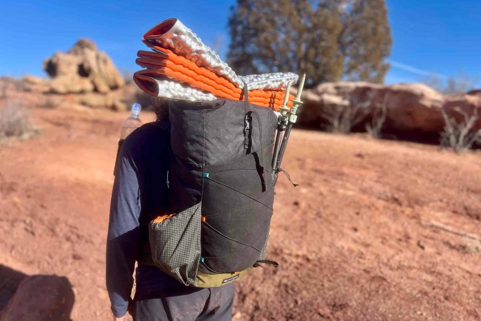 Back and side view of a black Atom Packs backpack with an orange and white sleeping pad strapped to the top and trekking poles secured to the side, showcasing the pack's external storage capabilities against a desert backdrop.