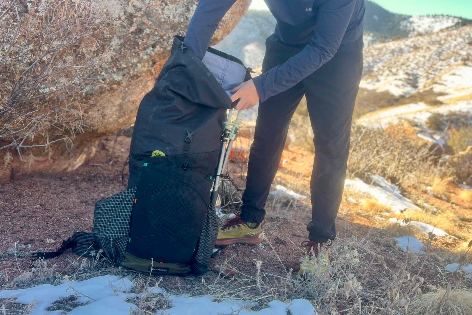 Side view of a black backpack with gridded mesh pockets resting on snowy ground, with hands adjusting trekking poles attached to the pack's side.