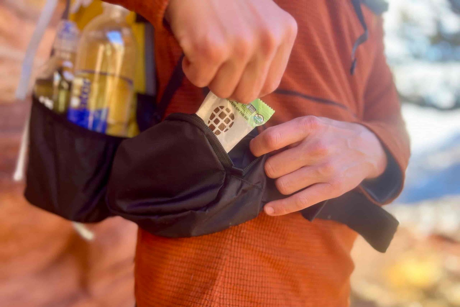 A person pulls a snack bar from the hip belt pocket of a backpack he is wearing.