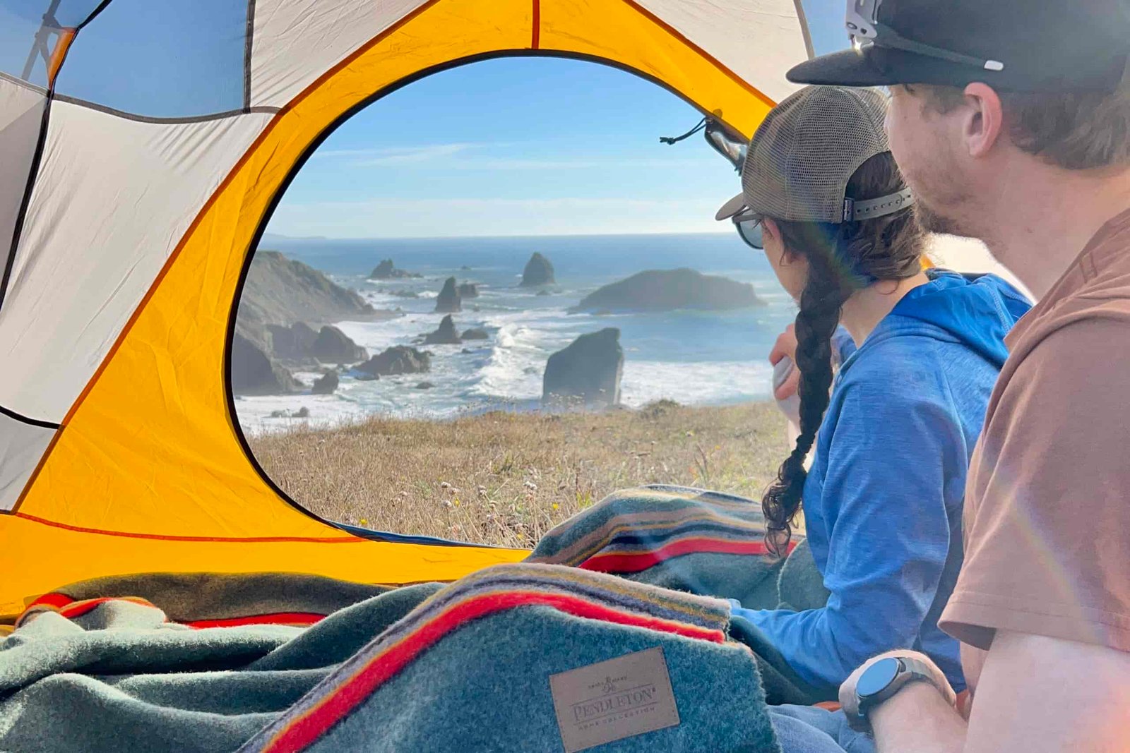 2 people sit inside of a tent overlooking the ocean while covered with the Pendleton Yakima Camp Blanket.