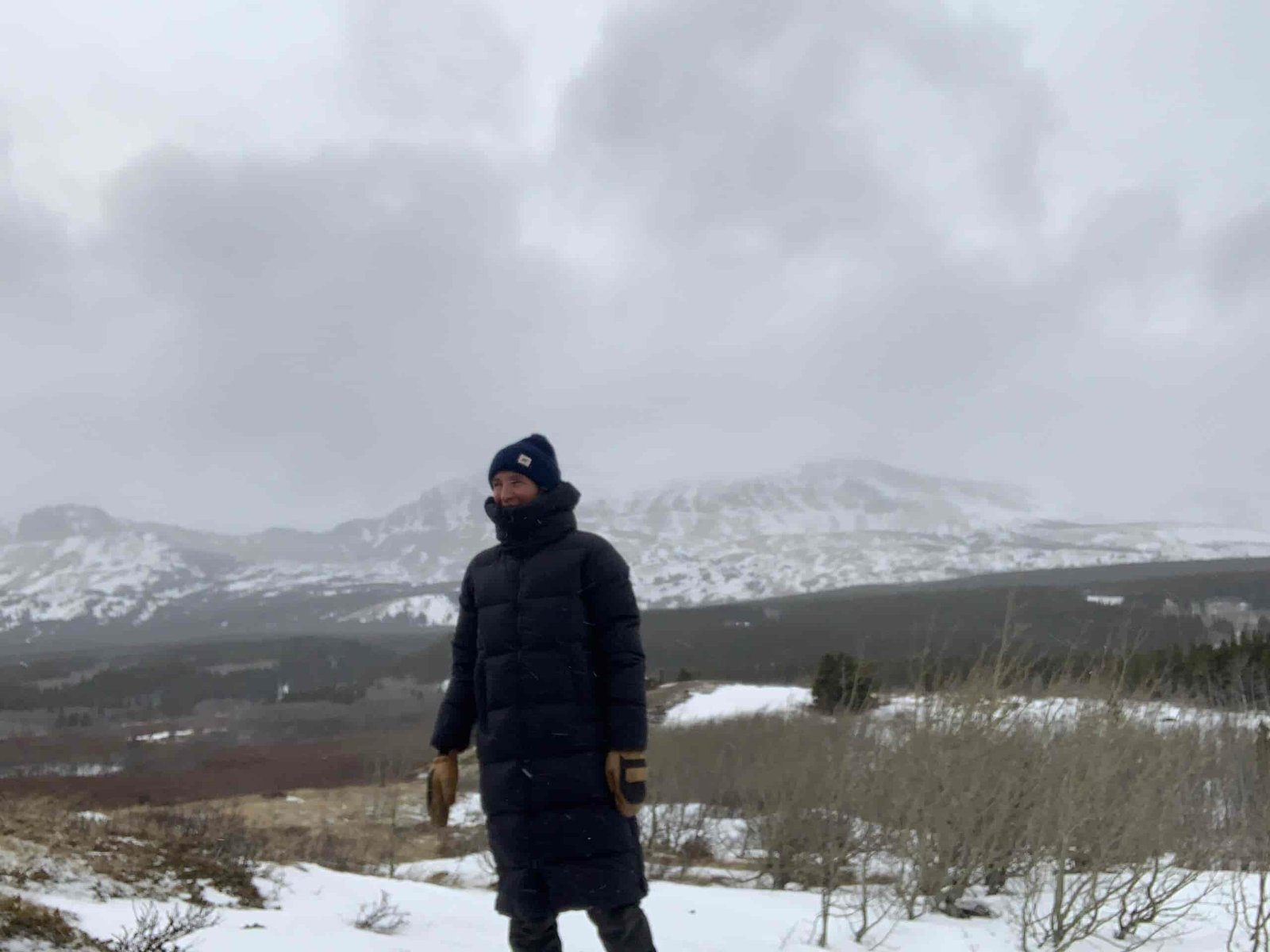 a woman standing below snow covered mountains on a windy pass while testing the quality of the outdoor research coze down winter jacket