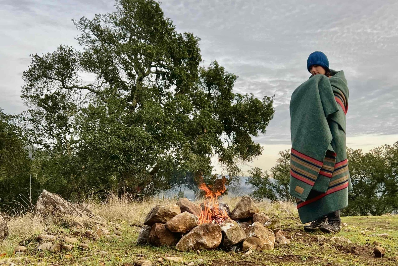 Women stands wrapped in the Pendleton Yakima Camp Blanket over a small campfire.