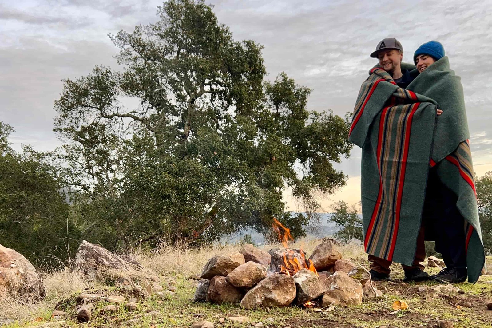 2 people stand wrapped in the Pendleton Yakima Camp Blanket near a cozy campfire.