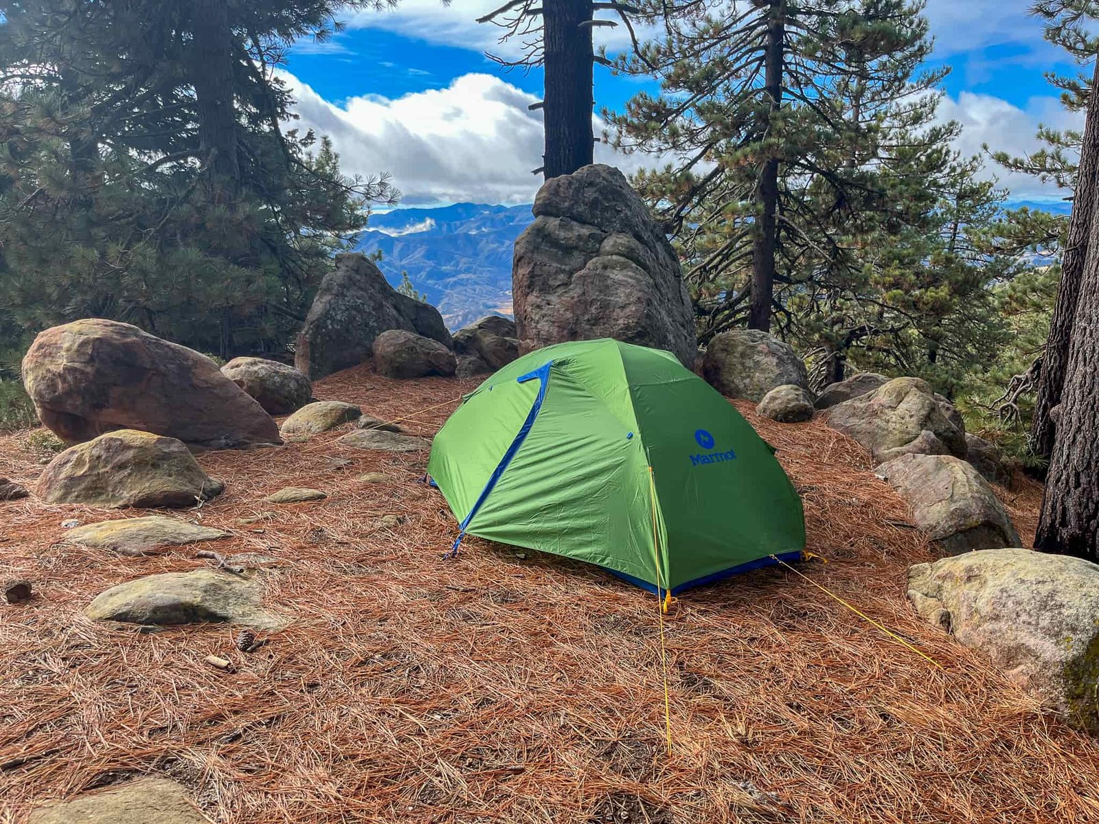 The Marmot Tungsten 2 set up on a bed of pine needles in a mountain clearing. Surrounded by large boulders and pines, with a distant view of mountain peaks in the background.