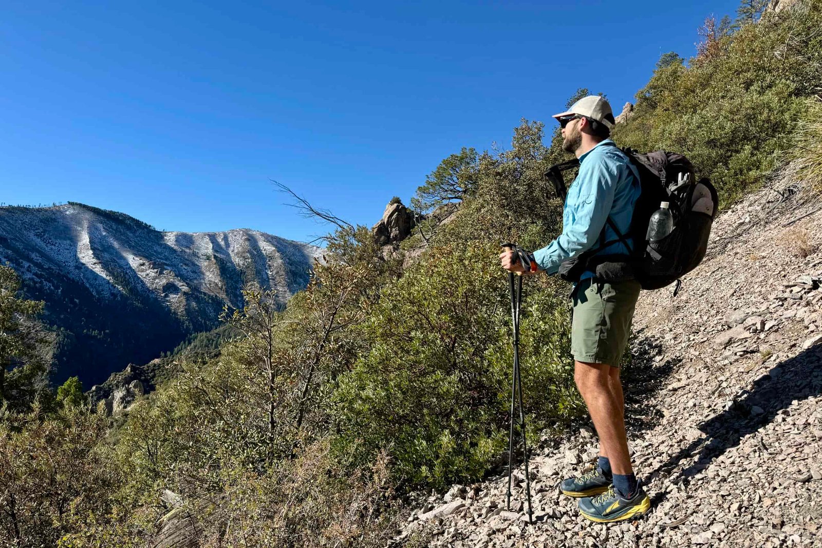 A person wearing a backpack and holding trekking poles is standing on a trail looking at mountain views.