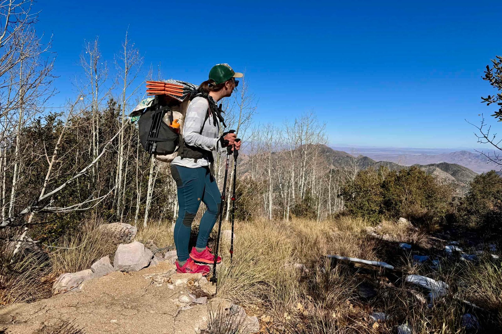 A person with a backpack is standing on top of a mountain and holding some trekking poles while looking out at a view.