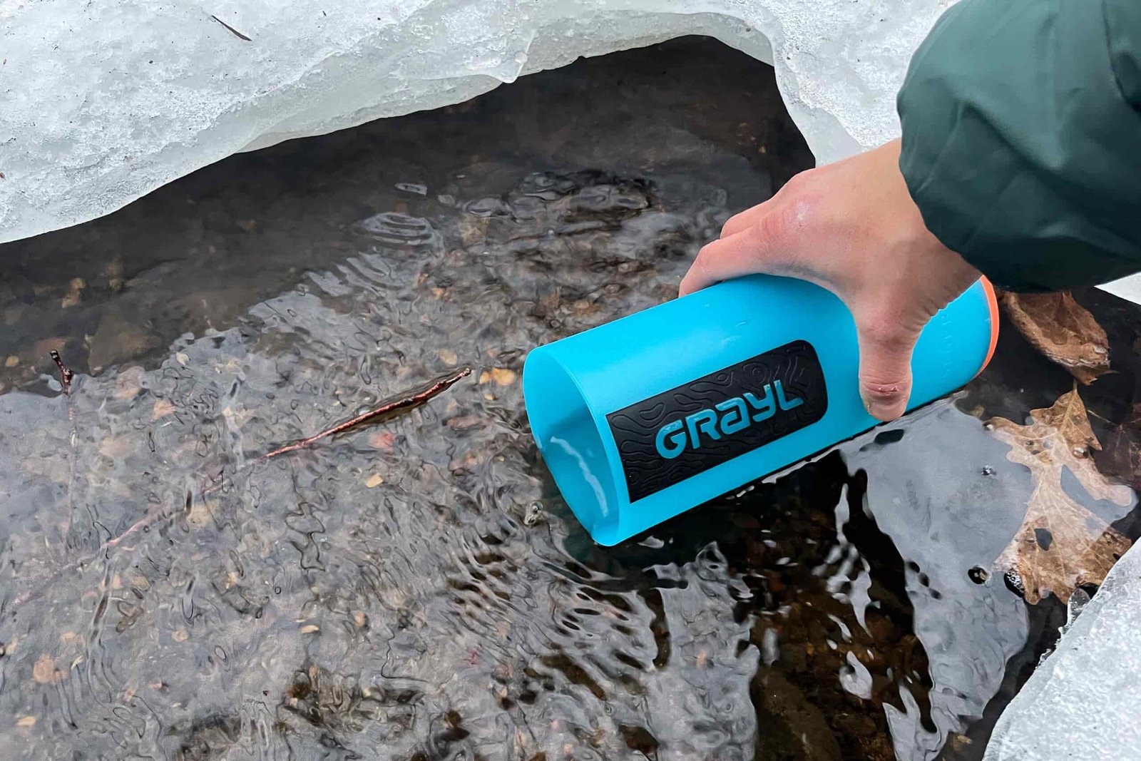 A close up of a person's hand dipping the Grayl GeoPress water bottle's outer cup into a partially frozen stream.