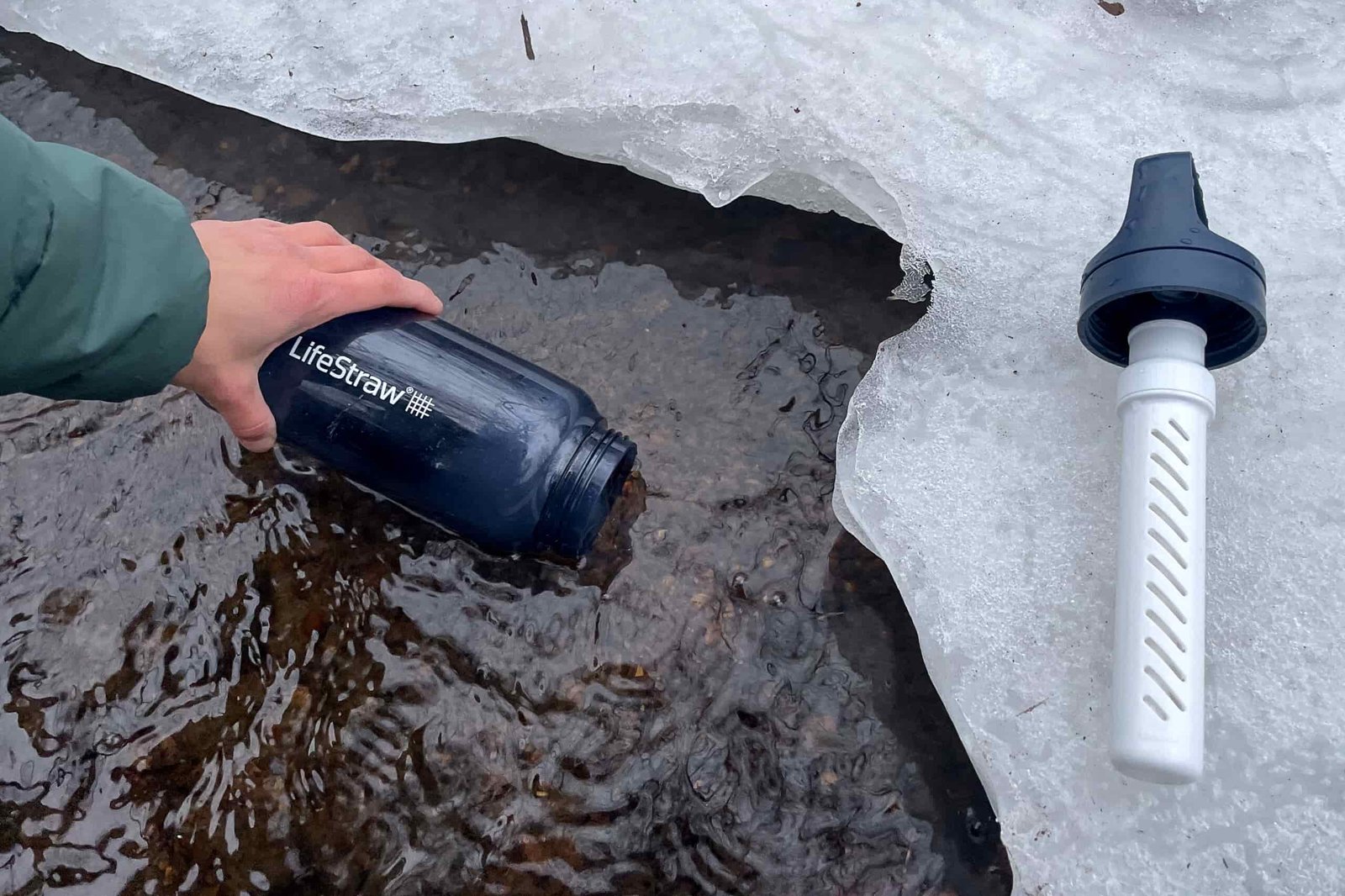 A person's hand holding the LifeStraw Go water bottle fills the bottle in a partially frozen stream. The bottle's lid and filter sit on the ice nearby.