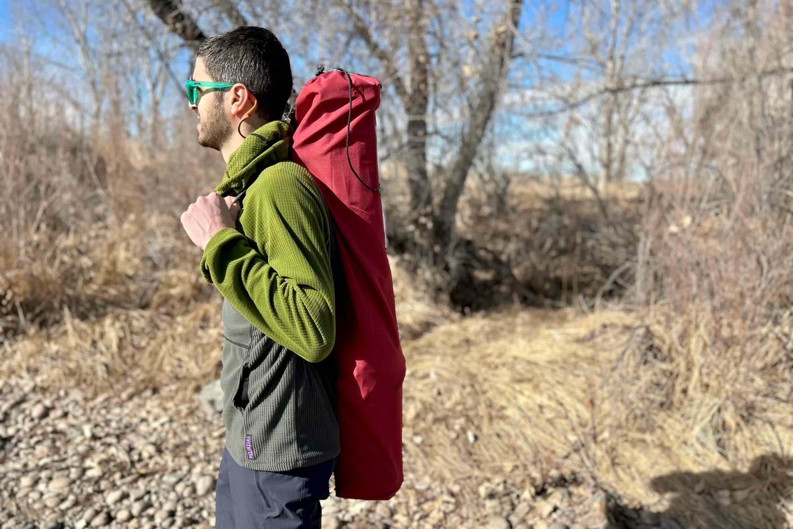 A man carries a camping chair in a carrying case on his back through the woods.