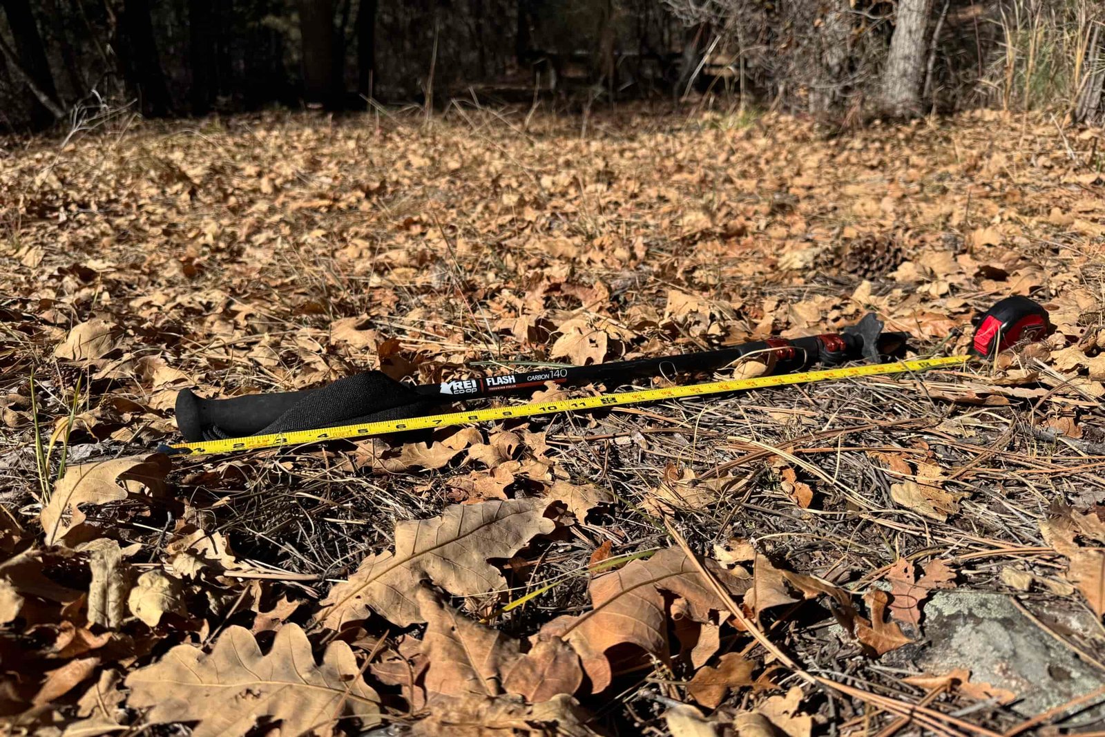 The REI Flash Carbon pole is laying on the ground in some leaves with a measuring tape next to it.