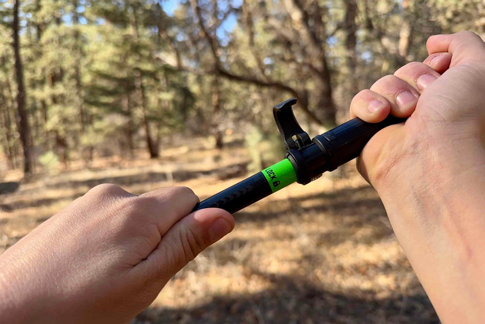 A close up of two hands pulling on a trekking pole to engage the tension lock on a pair of folding style poles. There is a forest in the background.