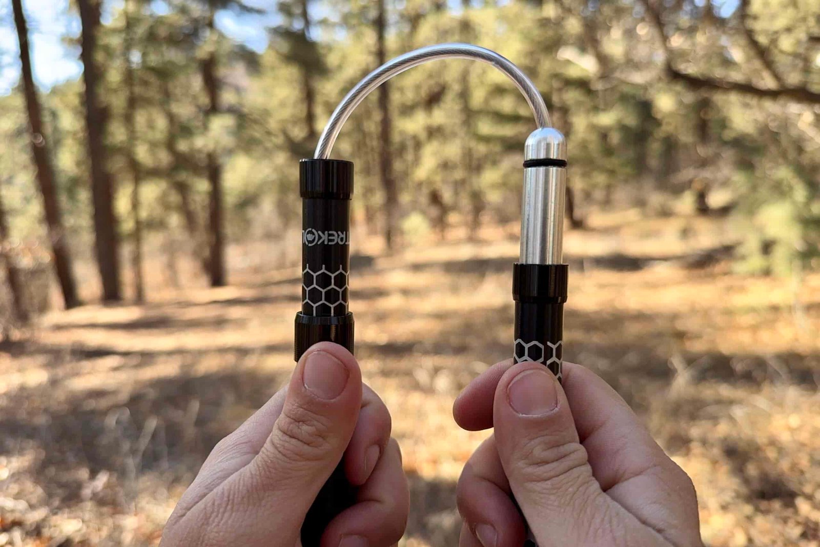 A close up of two hands each holding a shaft of a folding style pole. There is a cable cord connecting the shafts and a forest in the background.