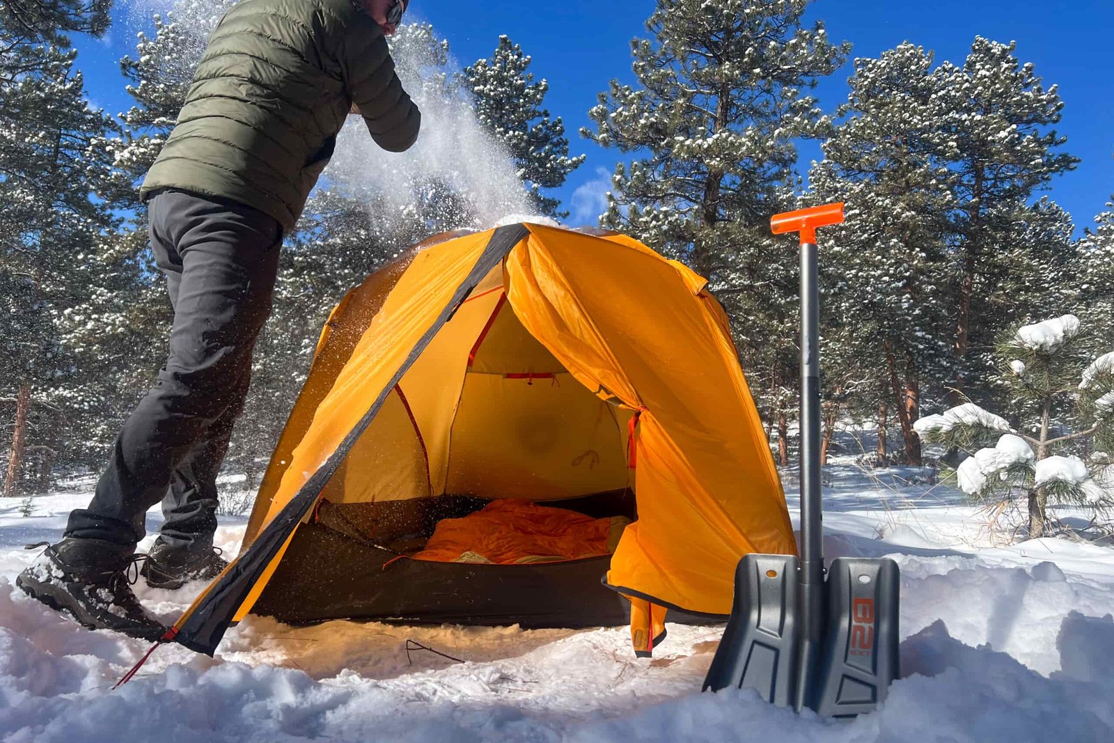 A camper brushing snow off a bright orange Nemo Kunai 2 tent under a clear blue sky, with a snow shovel standing upright in the foreground.