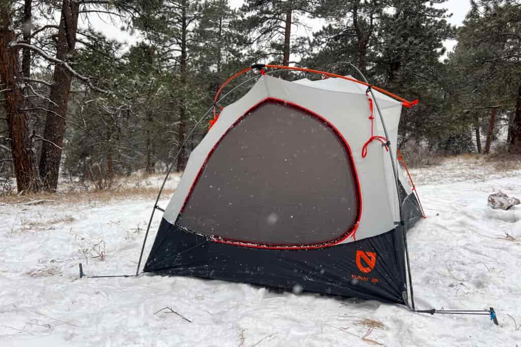 A Nemo Kunai 2P tent with a gray and black inner mesh layer pitched in a snowy forest, surrounded by pine trees with snow falling lightly.