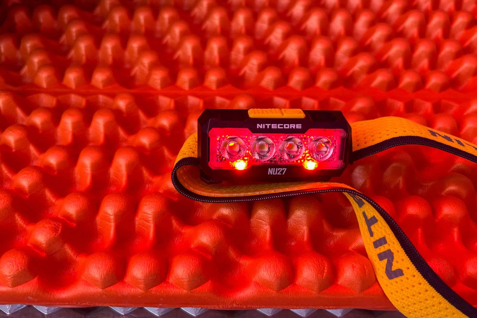 The Nitecore NU27 headlamp, with its red light function activated, shines directly at the camera while resting on a red foam sleeping pad.