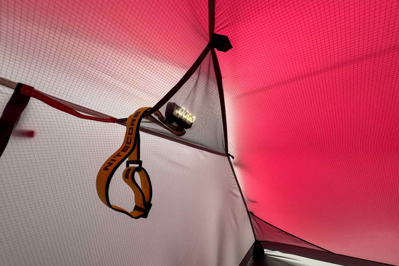 Nitecore NU27 headlamp hanging inside a tent, with its bright light illuminating the tent’s red and white interior while the detachable strap dangles below.