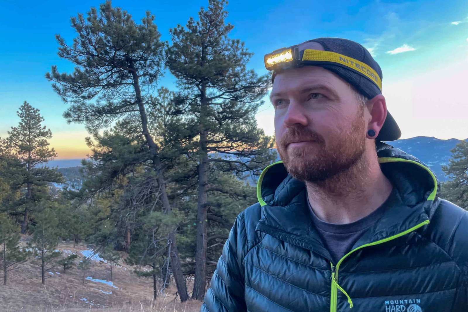 A man looking past the camera with a slight smile on his face. He's wearing a headlamp and the sun is setting in the background.
