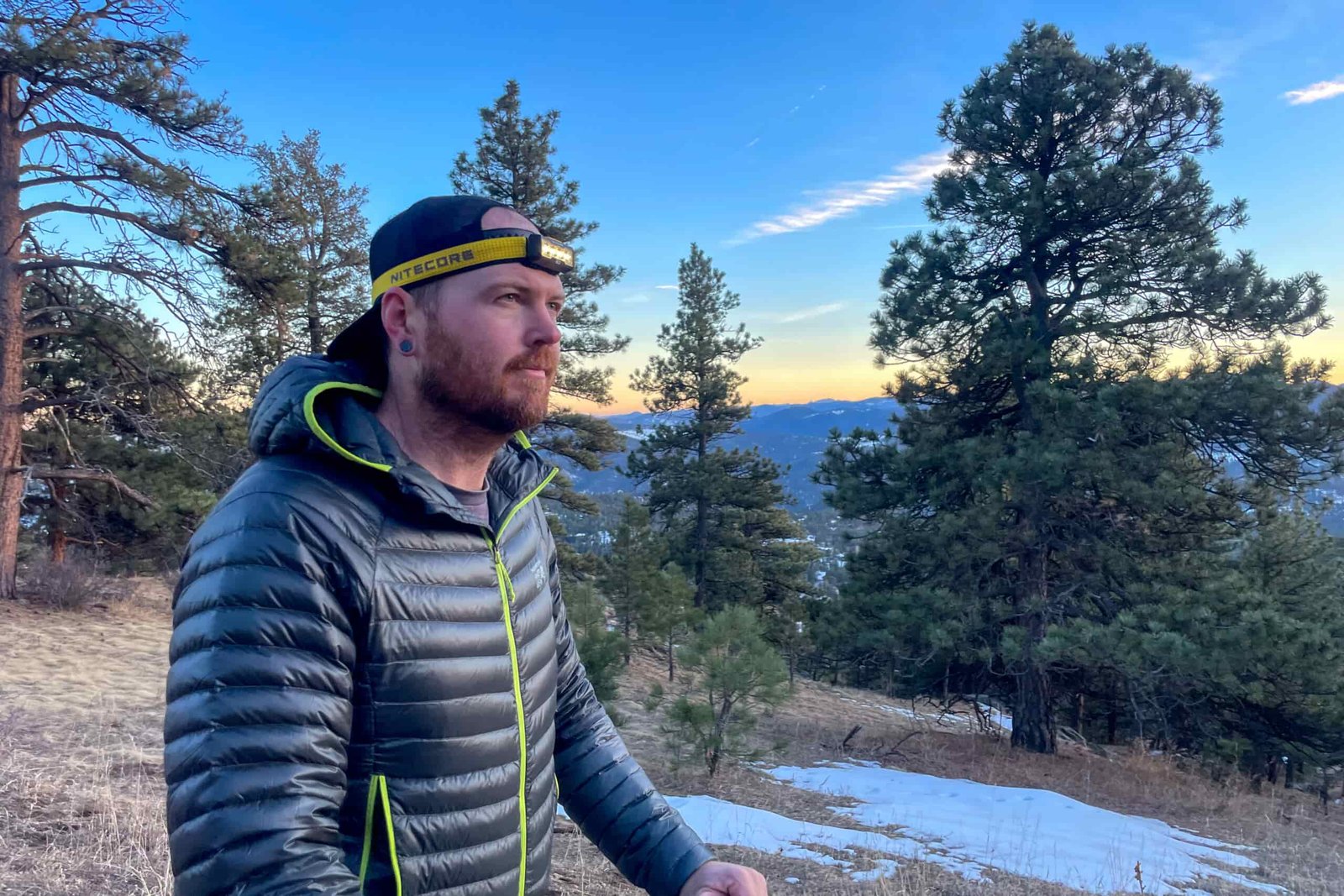 A man taking a hike in the mountains with the sun setting in the background. The man is hiking in colder weather and there are snow patches in the background.