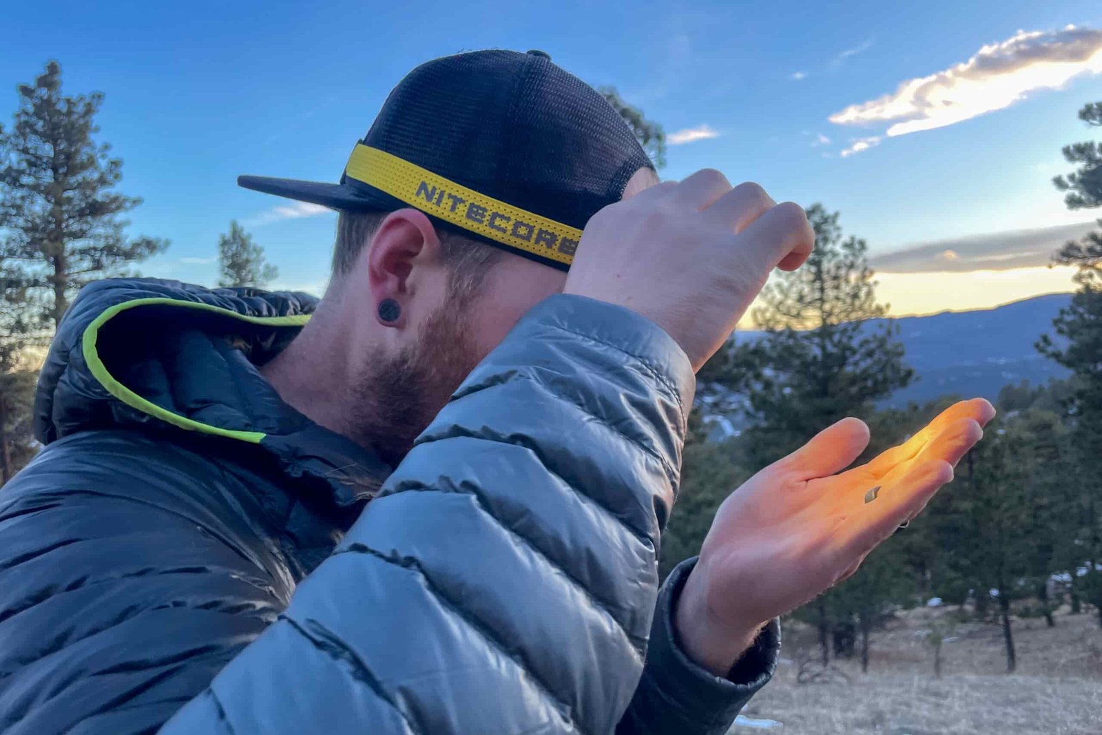 A man holds his hand in front of his to check the brightness of his headlamps. The Sun is setting in the background.