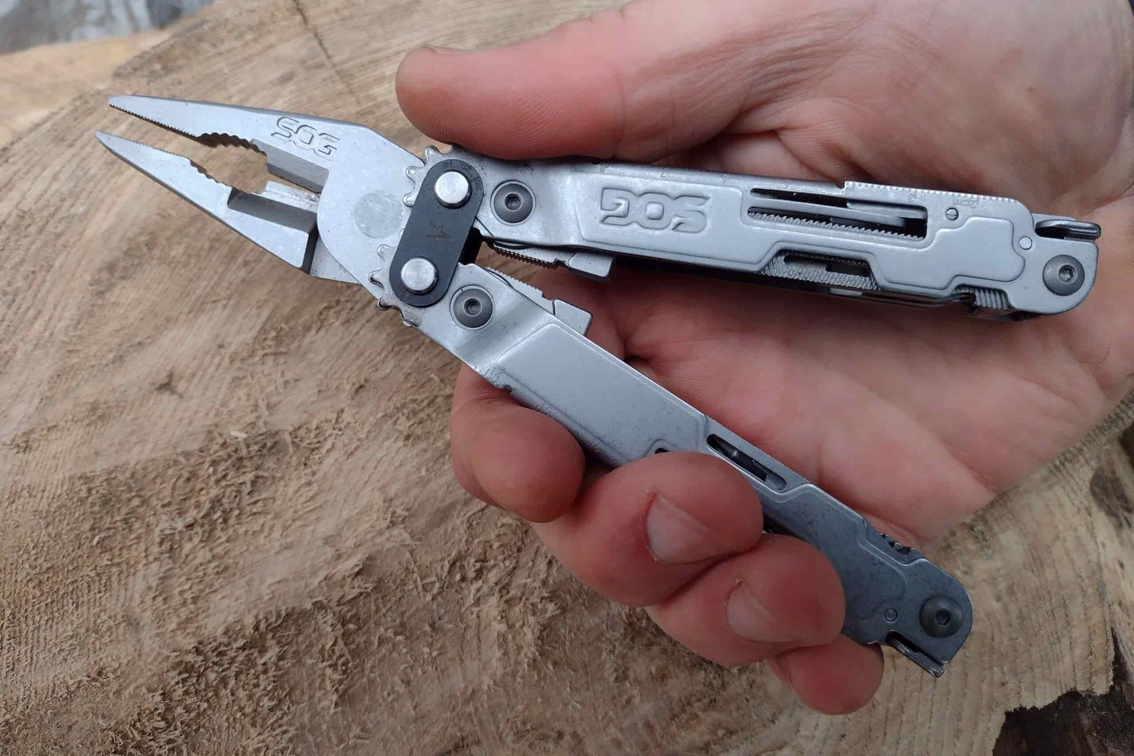 The silver SOG PowerAccess in hand, held above a log, with its needle nose pliers extended.