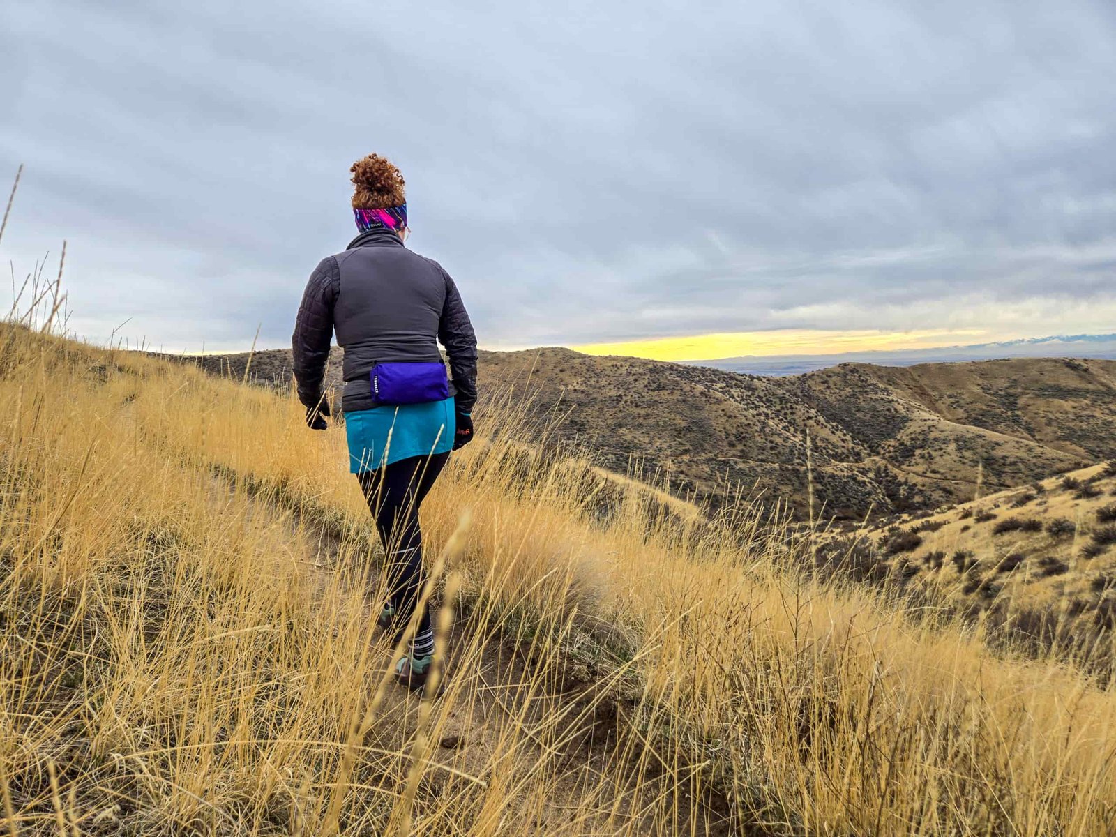 woman walking on a trail during sunset with dry grasses all around her and the hillsides