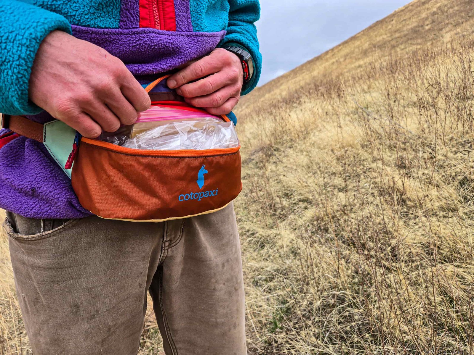 a man pulling a ziplock bag from his cotopaxi bataan fanny pack on a desert hillside