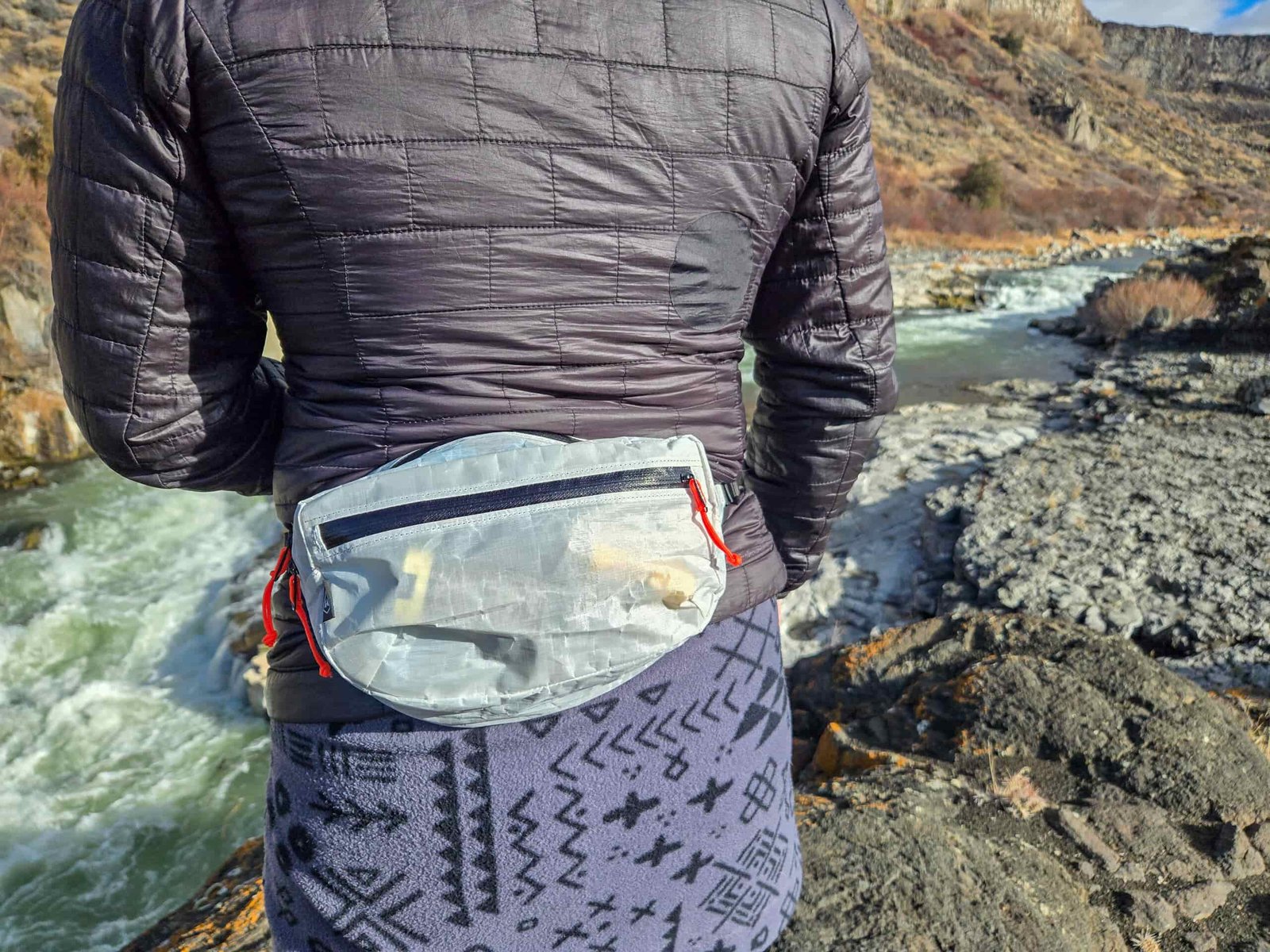 a woman wearing a hyperlite mountain gear versa fanny pack on her low back next to a rushing river
