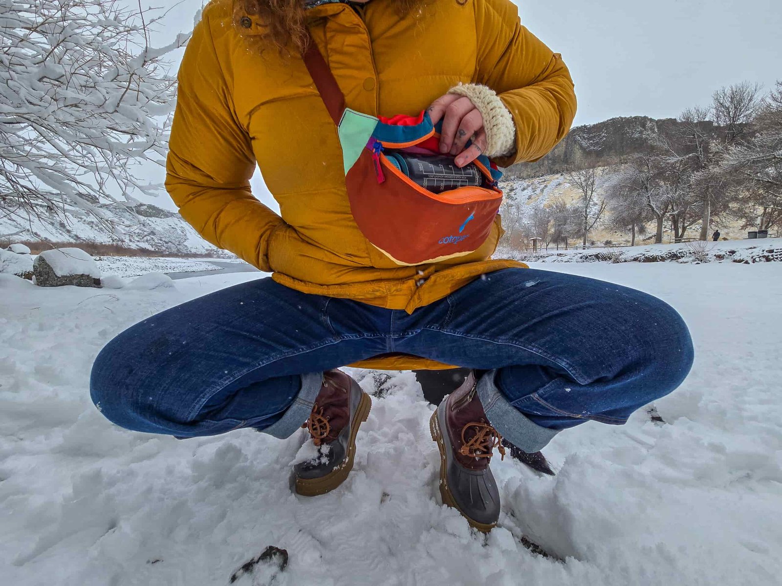 woman crouching and unzipping her cotopaxi fanny pack in a snowy park
