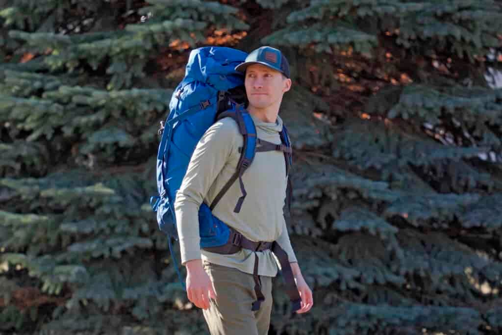 man wears blue backpack in front of a pine tree