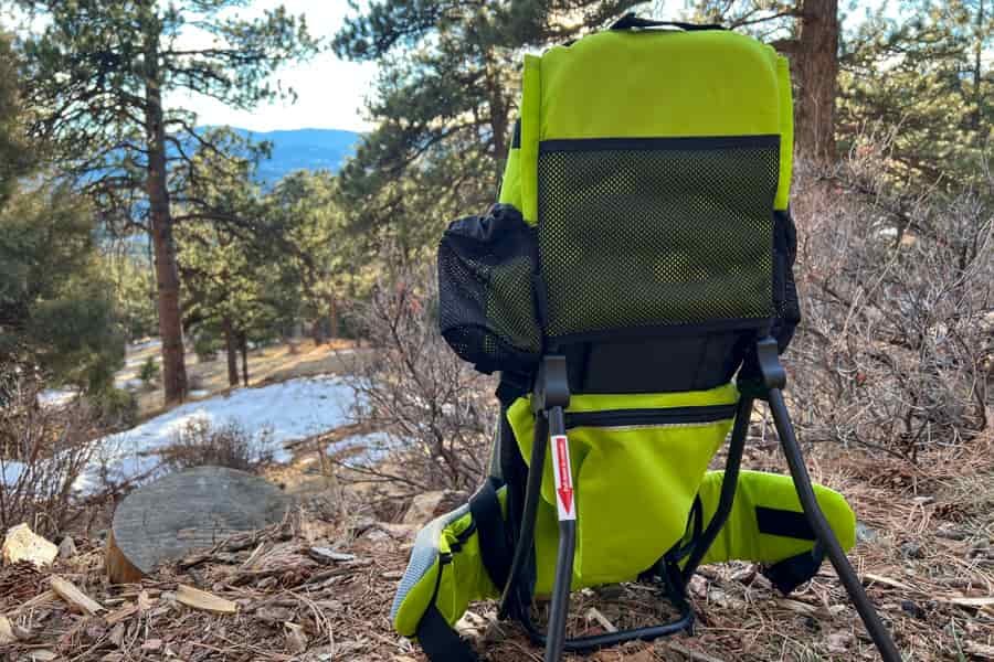 Rear view of a bright green child carrier against a mountain backdrop, showing its mesh back panel and storage compartments.