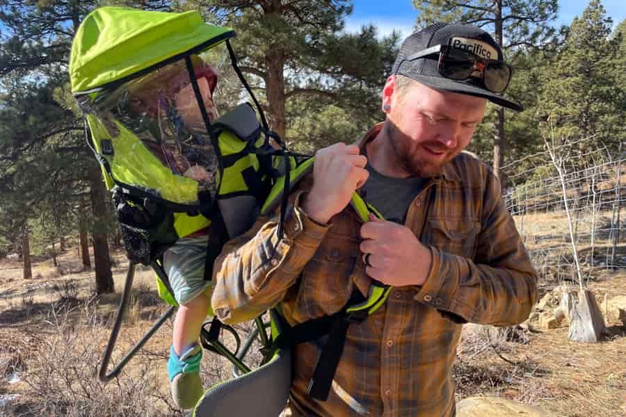 A man adjusting the straps of a bright green child carrier while carrying a child with a transparent rain cover outdoors.