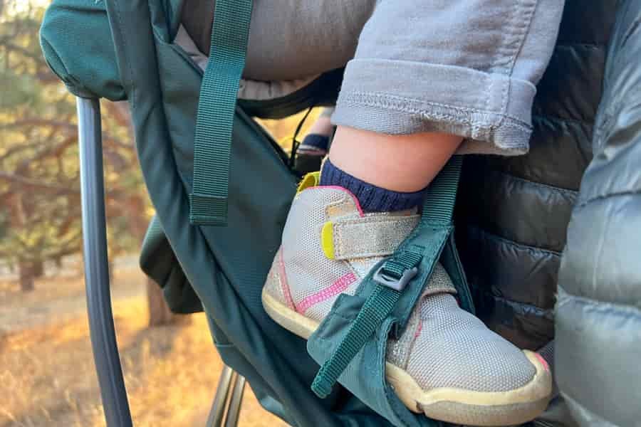 A close-up of a child's foot secured in the foot stirrup of a backpack carrier, highlighting the secure and adjustable straps.