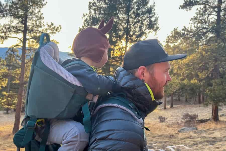 A side profile of the Deuter Kid Comfort carrier in use, showing the child’s seating position and supportive padding against a forest backdrop.