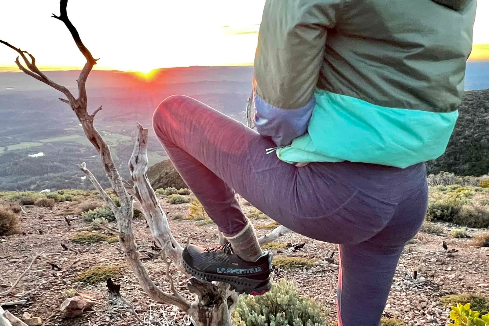 A woman stands watching the sunset with her foot placed on an old tree stump.