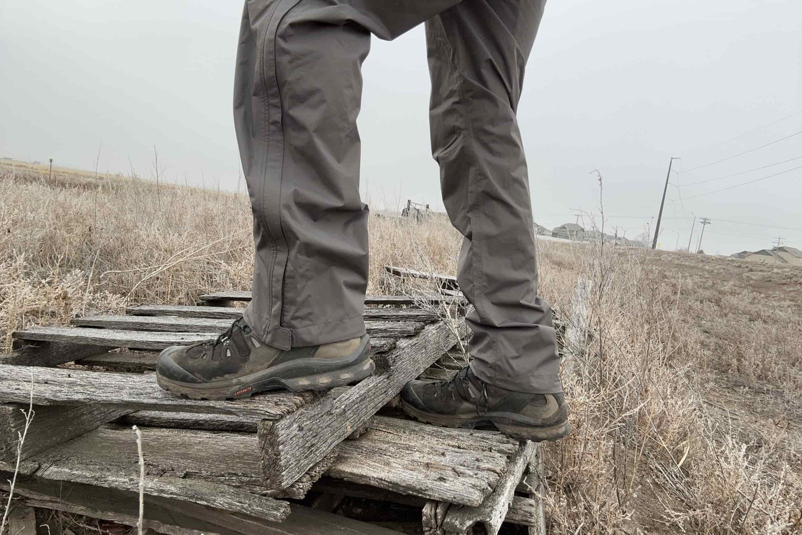 Closeup of someone wearing the REI Rainier rain pants, stepping up on old lumber in a field.