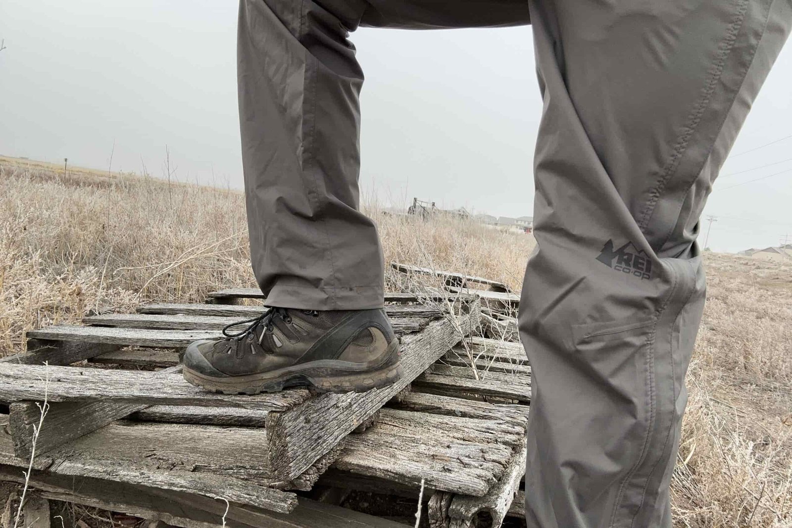 Closeup of someone wearing the REI Rainier rain pants, stepping up on old lumber in a field.
