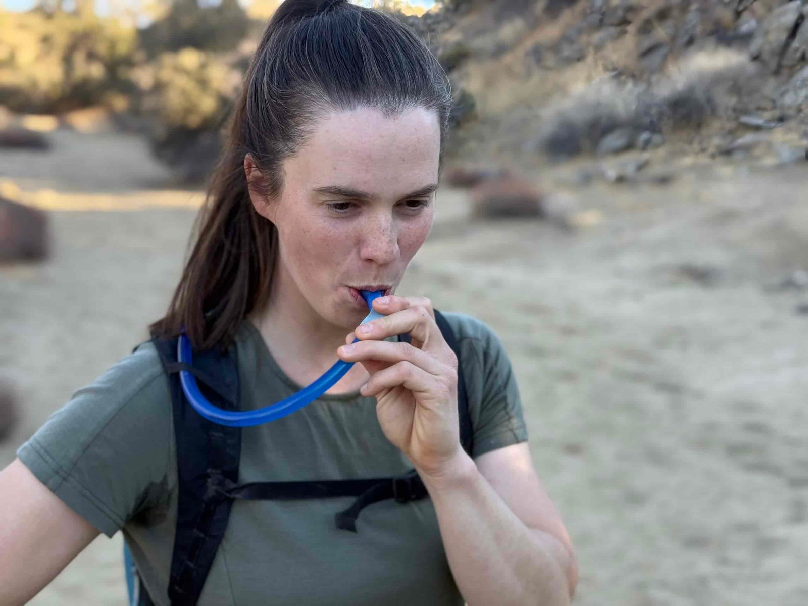 Hiker drinks from a hydration bladder standing in a sandy wash.