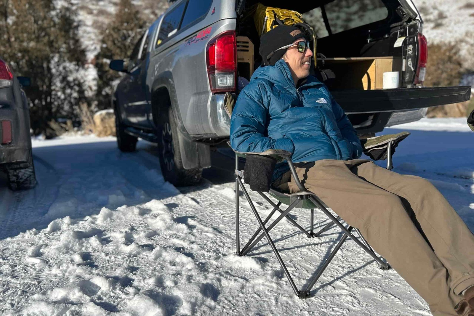 A man sits in a camping chair in a snowy parking lot while smiling.