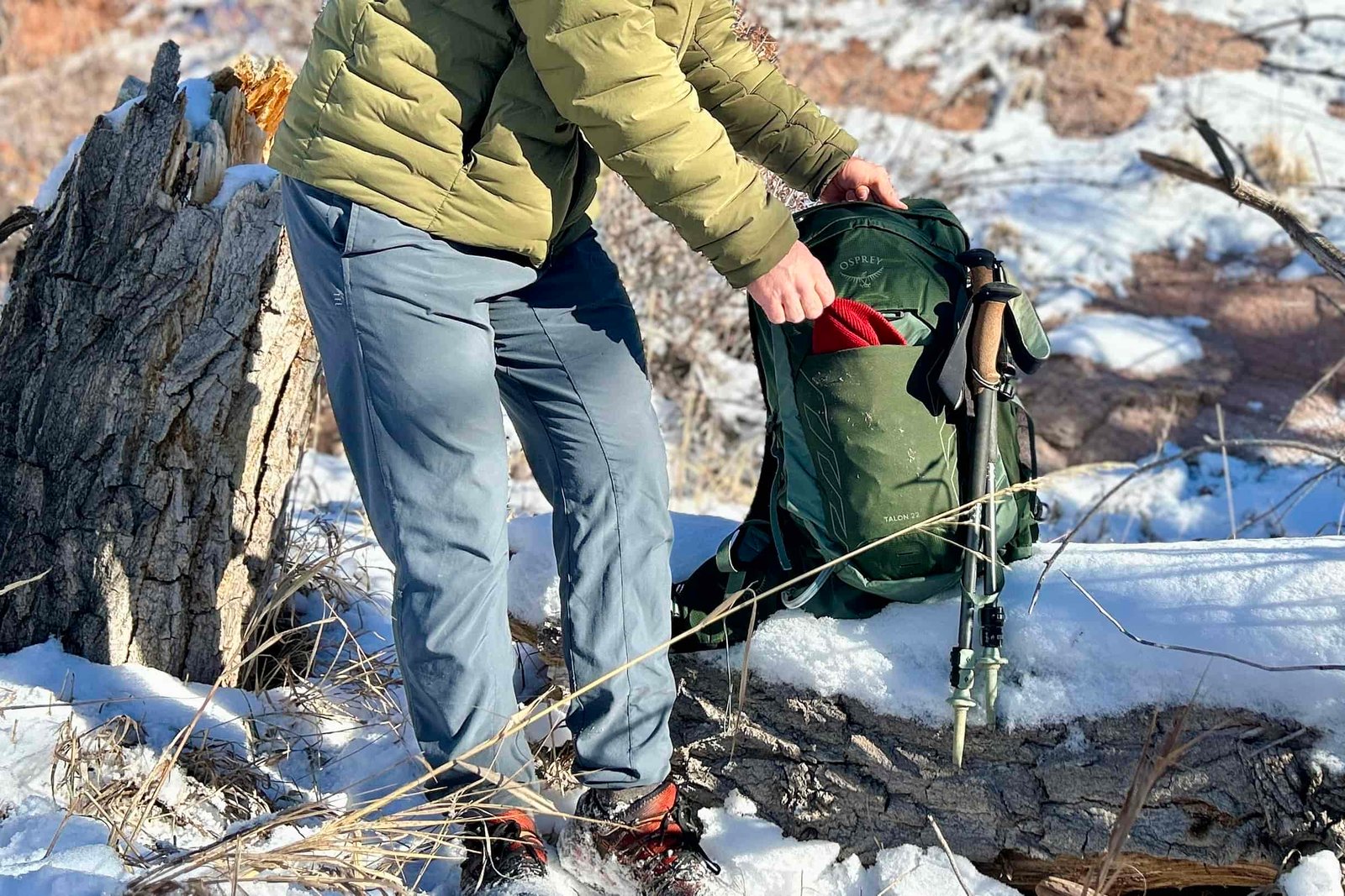 A person from the shoulders down pulling out a hat from their backpack that rests on a snowy log.