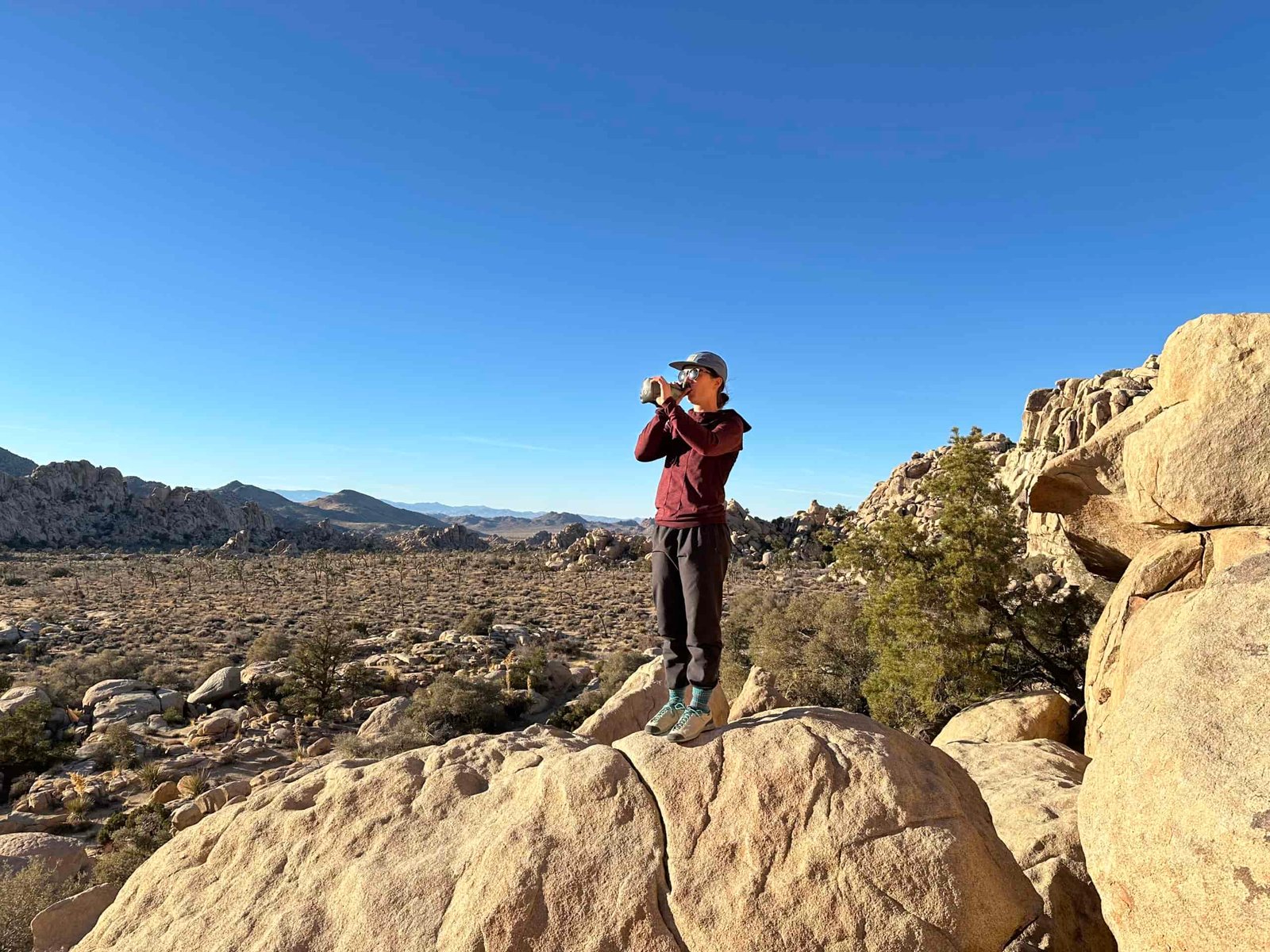 A far away shot of a hiker standing on a granite boulder while drinking out of the HydraPak Seeker water bag. The background shows the granite rock formations and scrub oak trees of Joshua Tree National Park.