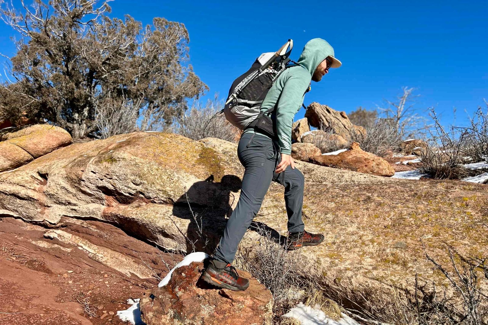 A man wearing a hoodie and a backpack hikes over sandy rocks in a desert setting.