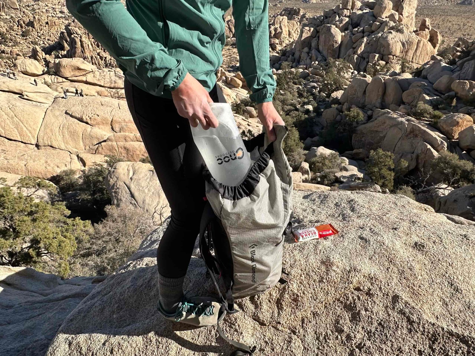 Close up of a hiker loading the CNOC VectoX into the top of her backpack. The background shows the granite rock formations of Joshua Tree National Park.