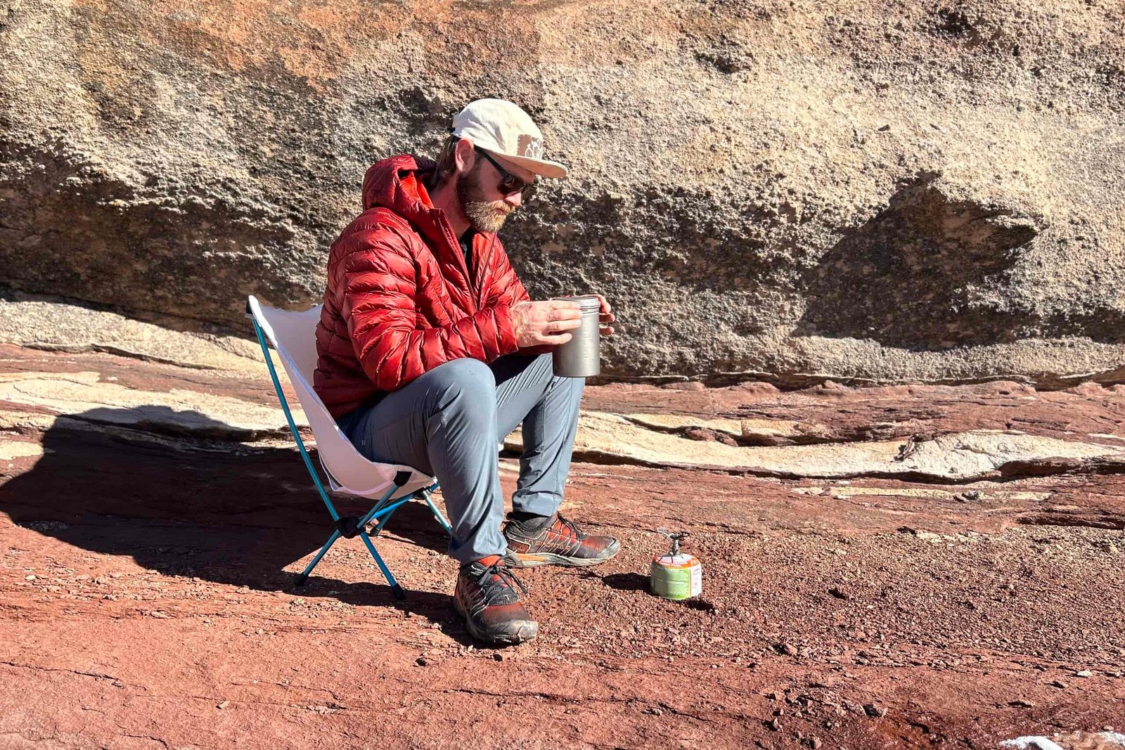 A man sits in a small camping chair holding a mug after cooking something on a small stove.