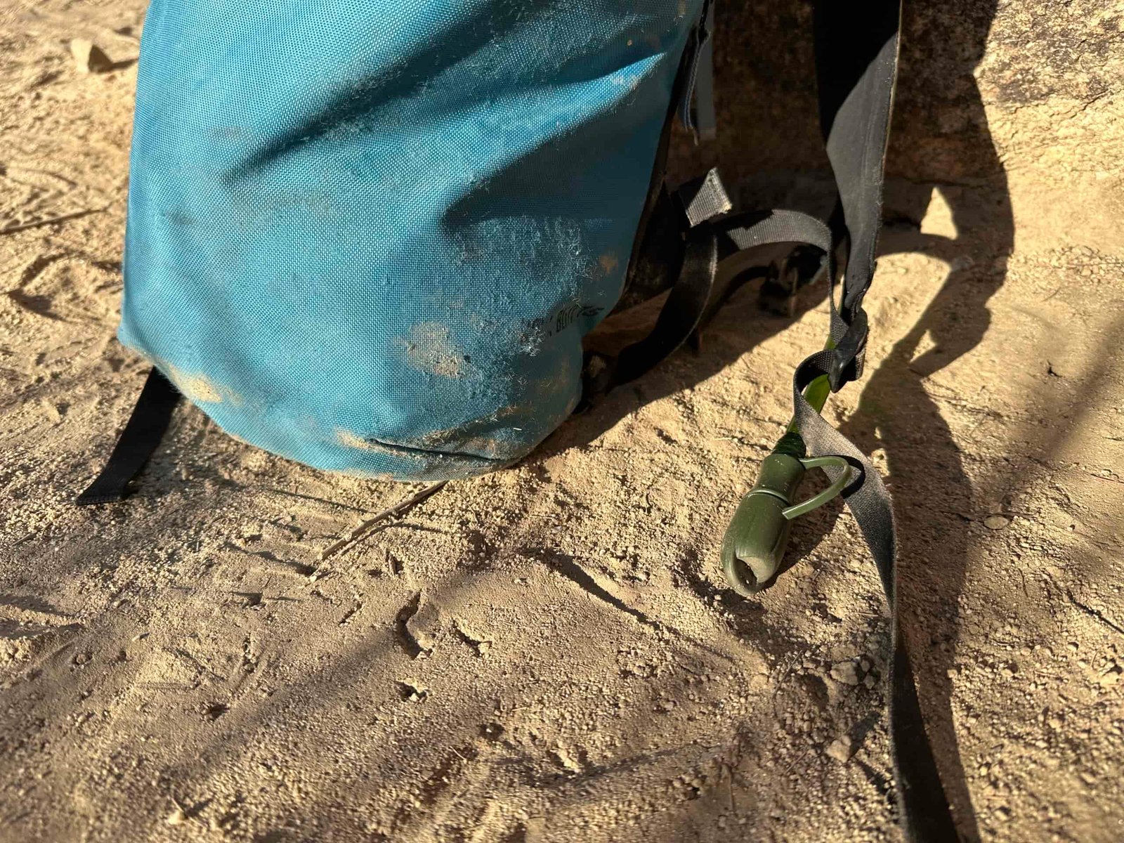 Close up of a hydration hose and bite valve coming out of a small backpack, all of which is sitting in the sand. The bite valve is protected with a cover.