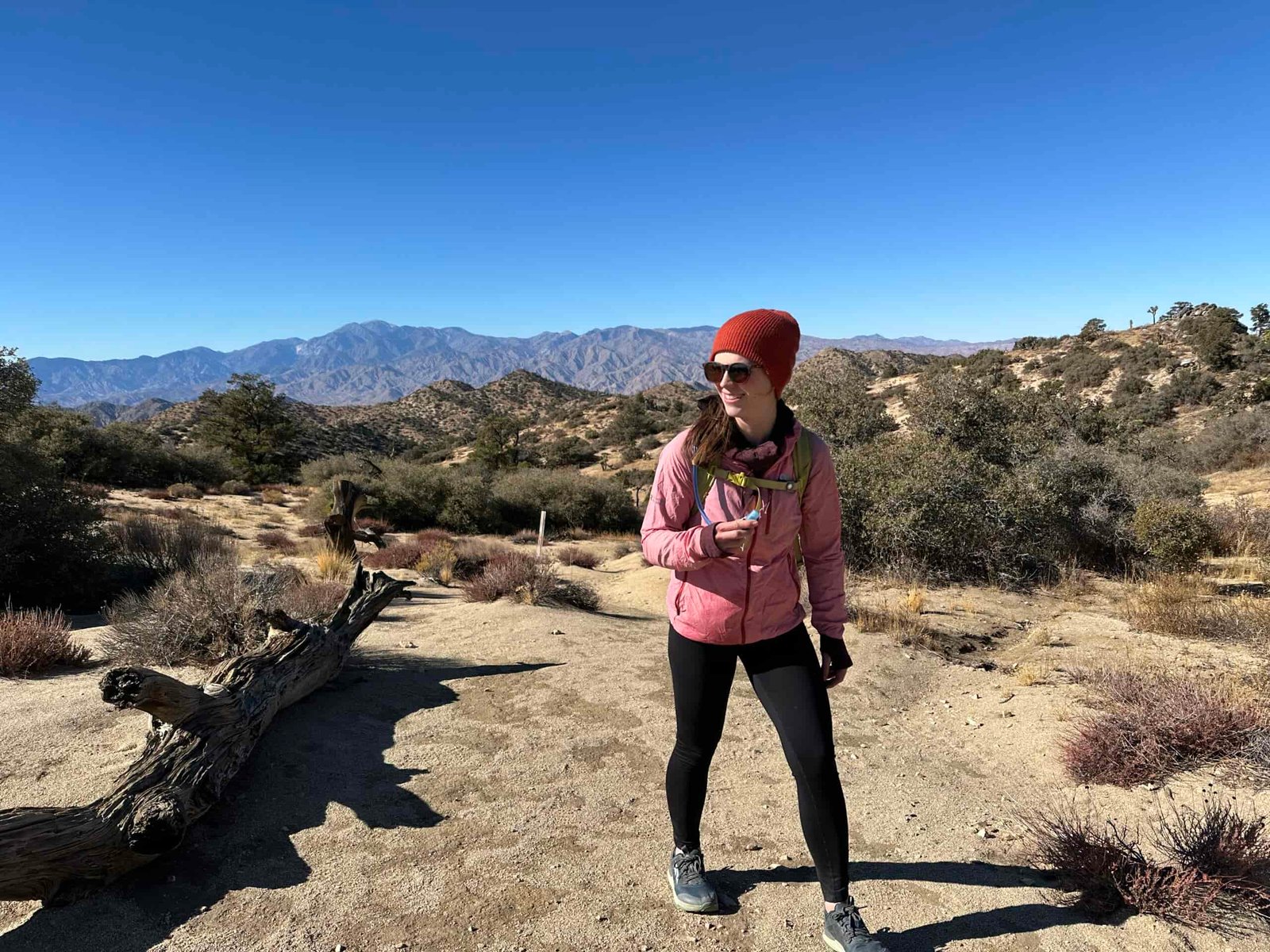 A hiker smiles while hiking on a trail in the Southern California desert. The trail is sandy with scrub oak bushes, and low, dry mountains are in the background.
