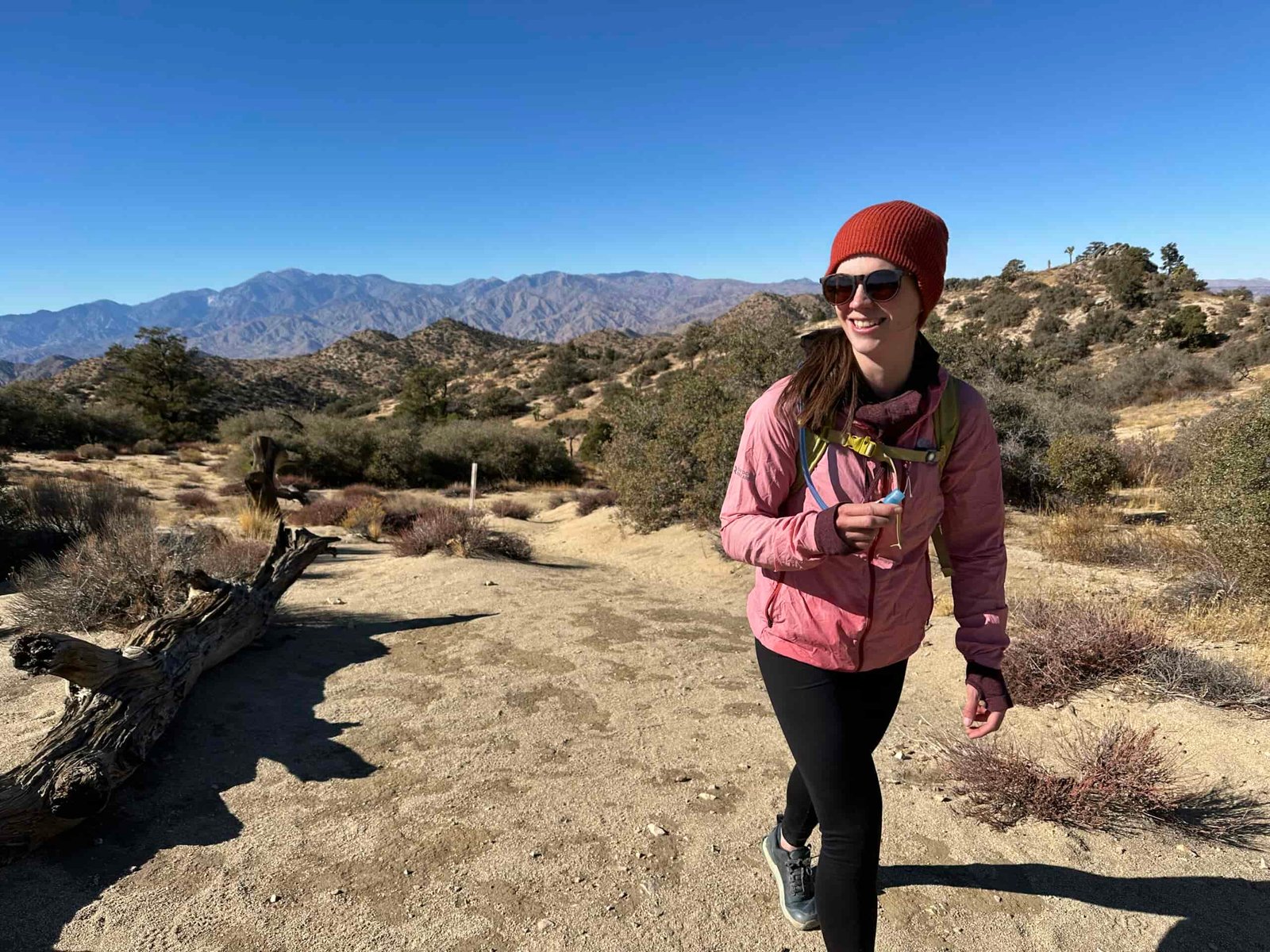 A hiker smiles while hiking on a trail in the Southern California desert. The trail is sandy with scrub oak bushes, and low, dry mountains are in the background.