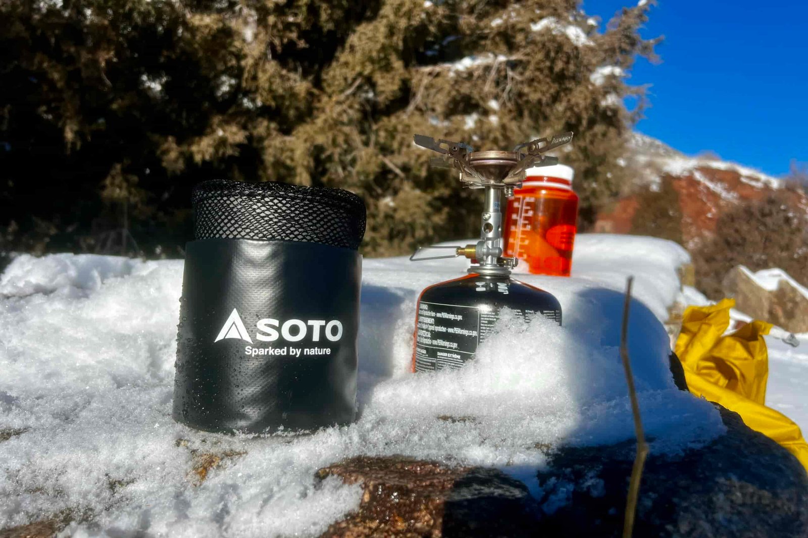 The Soto thermostack combo sits packed in its stuff sack on snow next to a stove and nalgene bottle with red rocks in the background.
