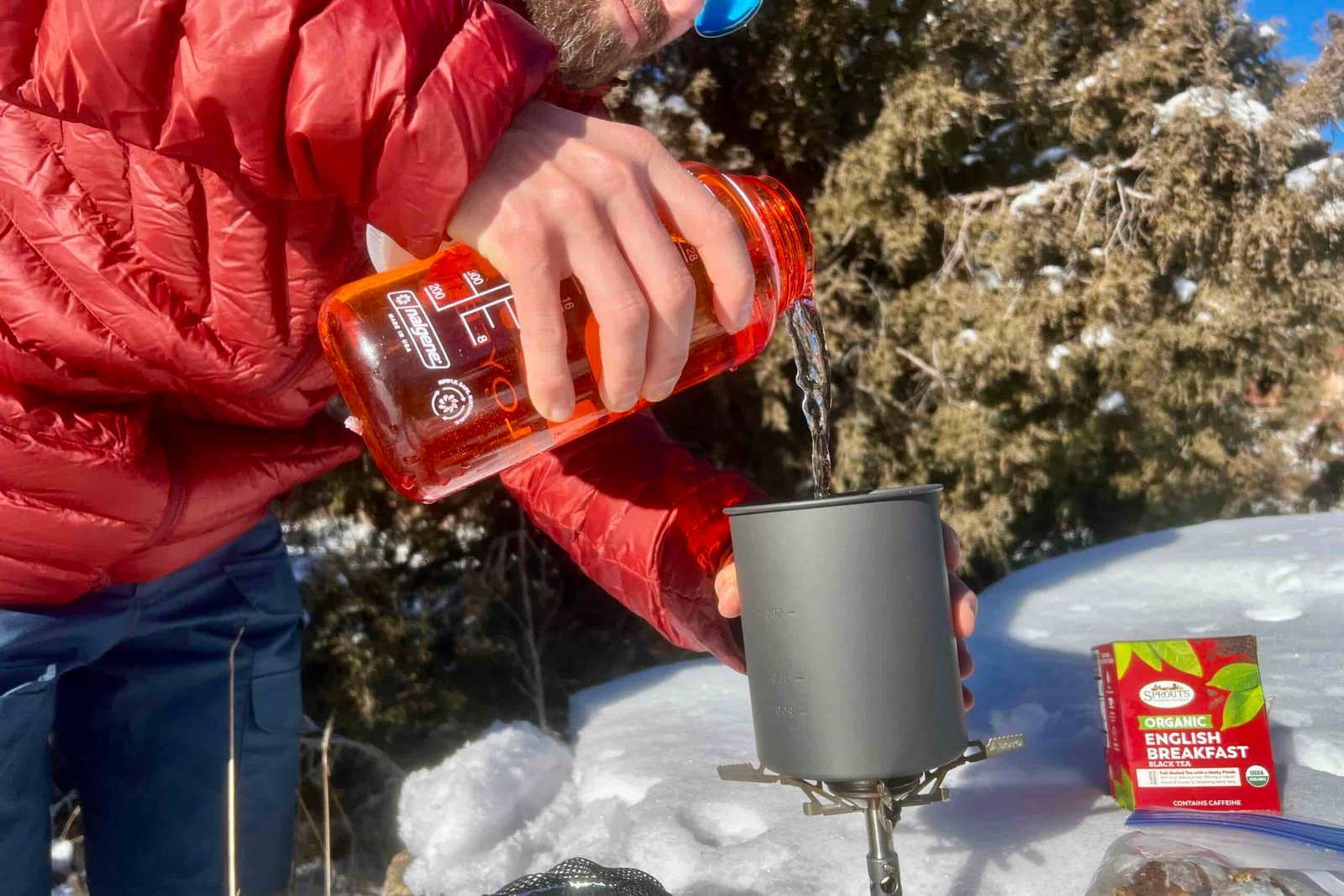 A hiker pours water from a nalgene bottle into the aluminum pot of the thermostack combo with tea nearby and a snowy background.