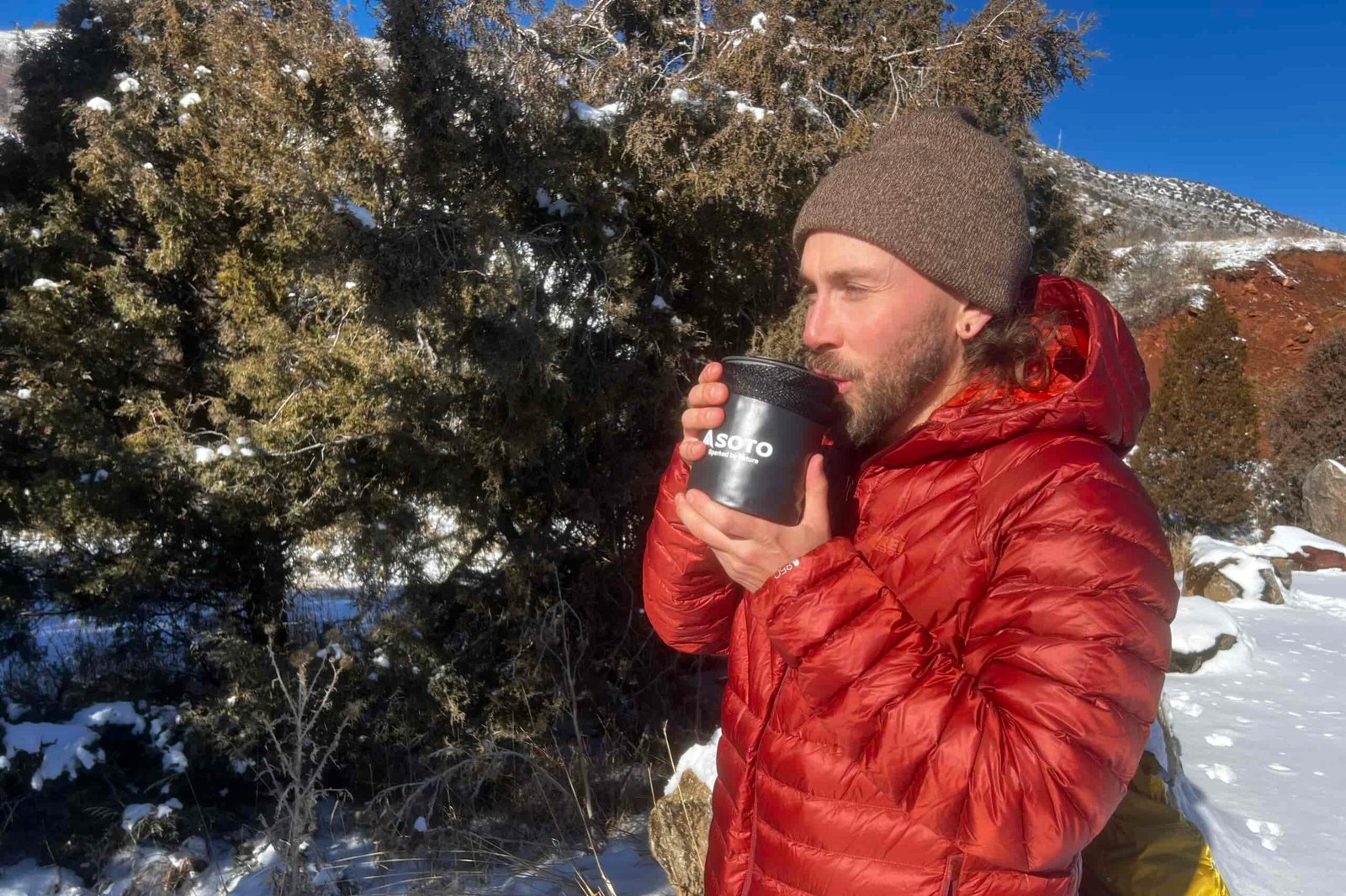A hiker sips from the insulated mug of the thermostack with it in the pot cozy. Snowy desert background.